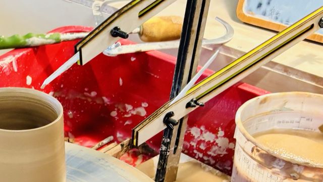 A wooden upright sitting in the basin of a pottery wheel with adjustable arms, one pointing at the rim of a freshly thrown mug. Each arm has a pointer at the end that can be flipped back out of the way.