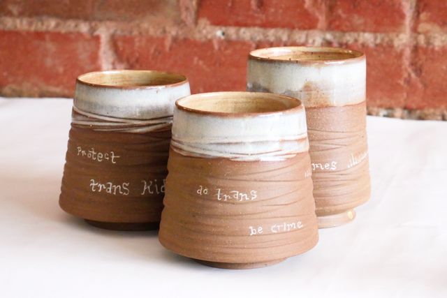 Three tumblers, also wide base with grooved walls sloped in - but the clay is brown un-speckled, the lips are an icy white, and they have hand-written text. The front one says "do trans, be crime". We can also see the end of "…crimes" sticking out on one behind, and "protect trans kids" on the last one
