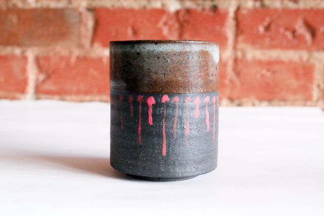 A small black tumbler, maybe 6oz, with red drippy-looking arrows pointing up to a grey-brown glazed rim
