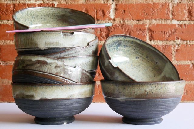 Two stacks of large black-clay ramen bowls with a deep foot and grooved texture around the bare clay base, then a frosty white glaze at the rim and inside -- and notches cut out for chopsticks.

