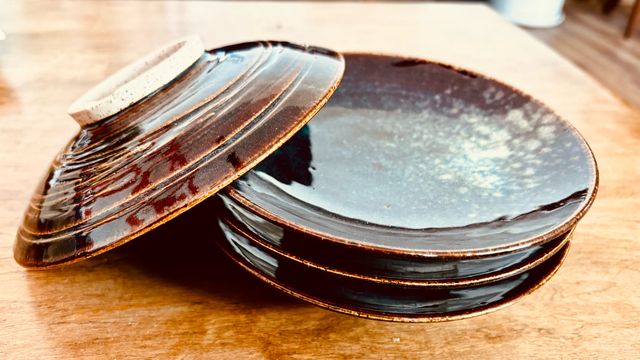A set of four shallow bowls stacked, with one overturned leaning against the others to show the deep foot ring and under-side texture. They have a deep glassy brown glaze with a splash of white and blue along one inside wall
