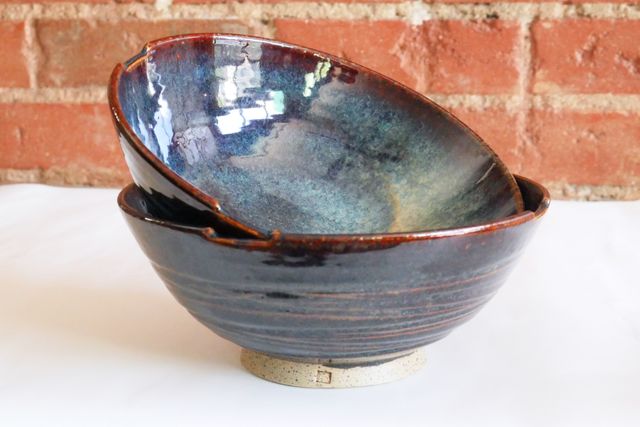 Two nearly identical soup bowls nested so we see the outer one from the side, a grooved texture that shows through the glassy dark brown-to-black-and iridescent blue. A rounded shape with a tall speckled clay foot ring and my small 'm' stamp. The inner bowl is off-kilter showing inside walls, smooth in the same glaze from a red-brown rim down into cosmic blues and a splash of yellow to one side.
