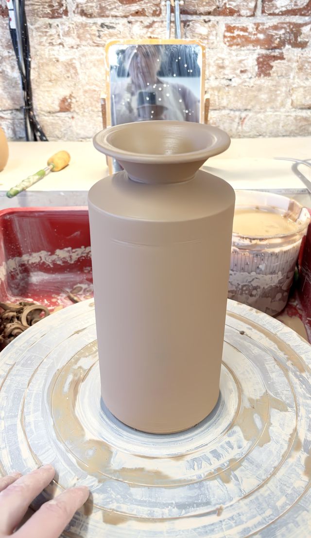 a tall cylindrical vase with straight walls up to a sharp angle in, then sharp angle back out, ending in a horizontal flared rim nearly the same diameter as the cylinder - reminiscent of an old-style milk-jug, but still wet on the wheel with splattered clay and tools around
