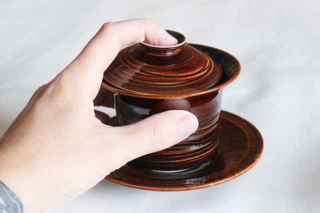 My hand holding a lidded gaiwan tea bowl just over the saucer - one finger pulling the lid back as though to pour tea strained through the small opening. All the parts are a glassy deep brown, breaking red over grooved textures, and the bowl has a wide flared rim for picking up when hot
