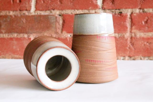 Two vases, one upright, the other on it's side so we can see into it - both conic like my mugs but taller, and with a rim that curves back down into the vase slightly to a narrower inset lip. A brown raw clay has deep grooves and little trails of red arrows, then a white rim at the top
