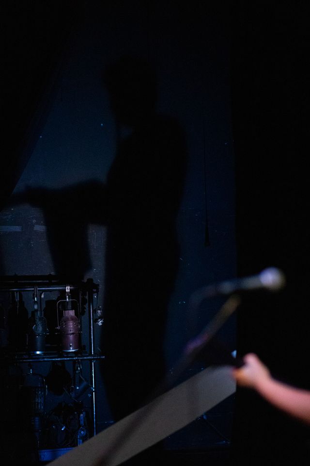 Josie's mic and hand in the foreground
but the focus is Miriam's shadow cast in blue light
on the back wall of the theater
next to a rack of stage lights
